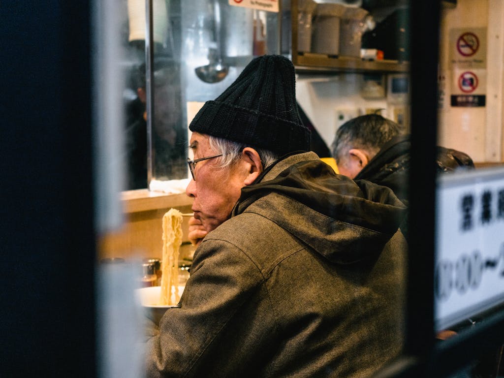 Side view of an elderly man eating ramen in a warm, cozy restaurant setting.